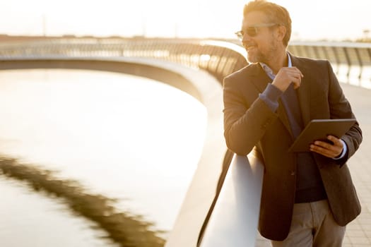 On a tranquil evening by the water, a professional man dressed in smart attire engages with his tablet, contemplating ideas while basking in the warm glow of sunset.