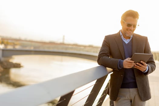 Wearing a stylish outfit, a man with a warm smile leans against a railing by the river at sunset, engaging with his tablet in a peaceful and picturesque environment.