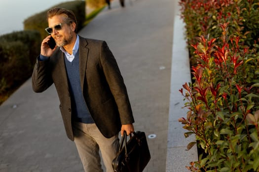 A confident businessman dressed in smart attire walks beside vibrant red plants, conversing on his mobile phone as the sun sets over the waterfront, reflecting a busy yet serene atmosphere.