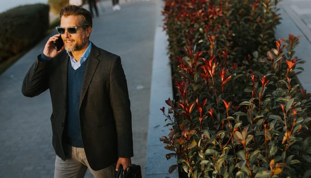A well-dressed man strolls along a riverside walkway while talking on his phone. The vibrant plants and calming water create a serene atmosphere perfect for business discussions.