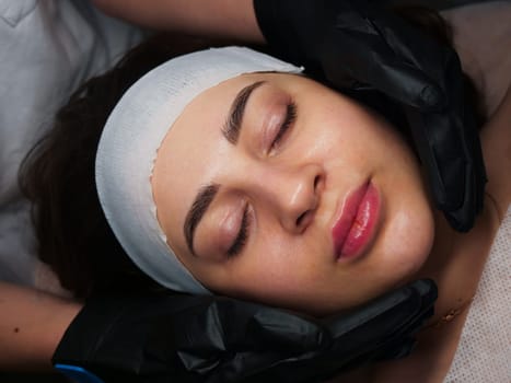 The beautician performs the procedure of microcurrents in black electric gloves to restore facial skin cells. Close-up