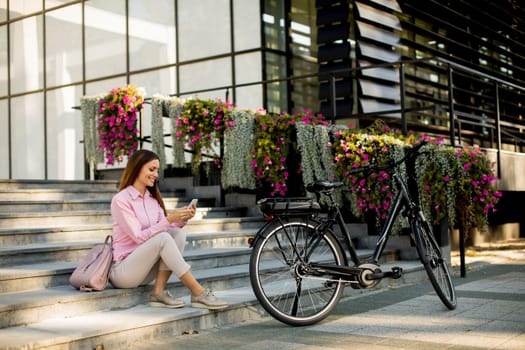 Young woman sitting on the stairs near ebike and usling smartphone outdoor