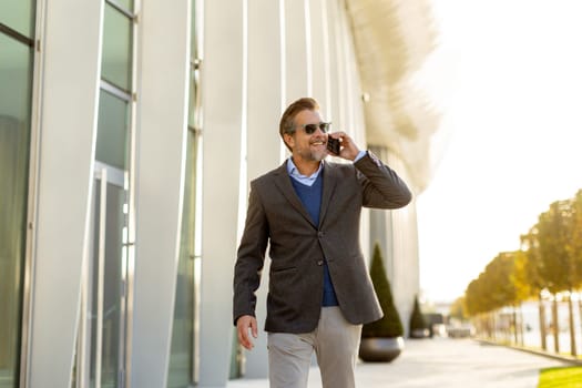 A well-dressed man strolls along a sleek pathway, chatting on his phone under the warm glow of the afternoon sun, surrounded by contemporary architecture and greenery.