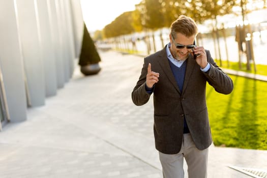 A well-dressed man strolls along a sleek pathway, chatting on his phone under the warm glow of the afternoon sun, surrounded by contemporary architecture and greenery.