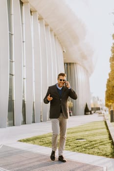 A well-dressed man strolls along a sleek pathway, chatting on his phone under the warm glow of the afternoon sun, surrounded by contemporary architecture and greenery.