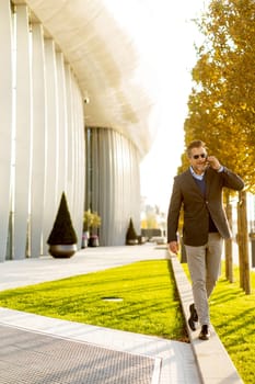 A well-dressed man strolls along a sleek pathway, chatting on his phone under the warm glow of the afternoon sun, surrounded by contemporary architecture and greenery.