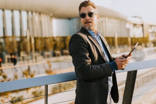 An elegant individual in a blazer stands on a balcony at sunset, holding a tablet and gazing over a vibrant metropolis, embodying professionalism and relaxation.