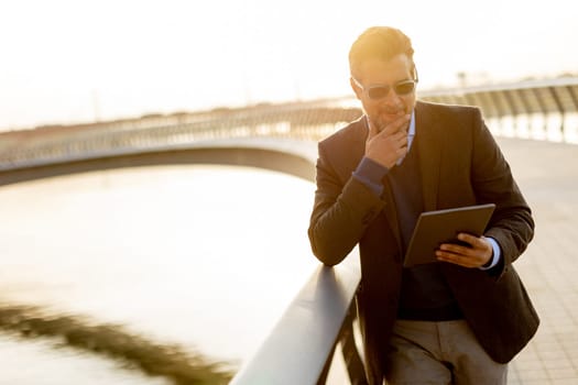 On a tranquil evening by the water, a professional man dressed in smart attire engages with his tablet, contemplating ideas while basking in the warm glow of sunset.