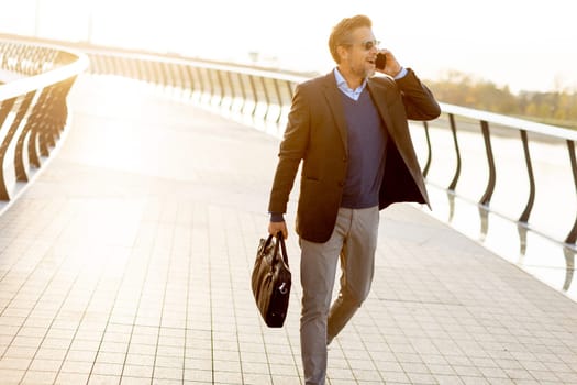 A well-dressed man walks confidently along a picturesque waterfront path, checking his phone, with colorful leaves and a serene backdrop accentuating the tranquil golden hour.
