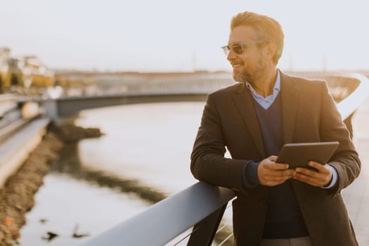 Wearing a stylish outfit, a man with a warm smile leans against a railing by the river at sunset, engaging with his tablet in a peaceful and picturesque environment.