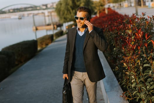 A well-dressed man strolls along a riverside walkway while talking on his phone. The vibrant plants and calming water create a serene atmosphere perfect for business discussions.