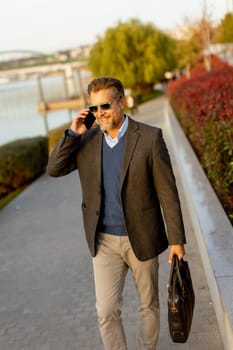 Dressed in a smart casual outfit, a man walks confidently by the water, engaged in a phone conversation while admiring the vibrant autumn colors around him