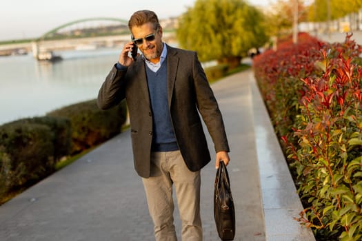 Dressed in a smart casual outfit, a man walks confidently by the water, engaged in a phone conversation while admiring the vibrant autumn colors around him