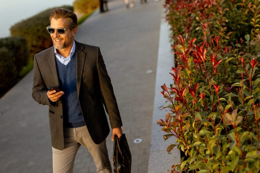 A well-dressed man walks confidently along a picturesque waterfront path, checking his phone, with colorful leaves and a serene backdrop accentuating the tranquil golden hour.