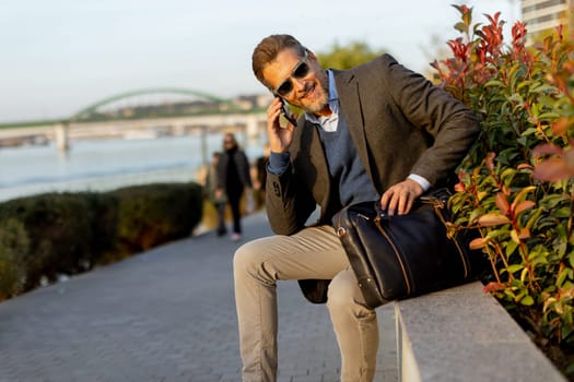 A professional man dressed in a blazer enjoys a sunny afternoon, chatting on his phone beside lush plants while carrying a leather bag. The scene reflects a blend of work and relaxation.