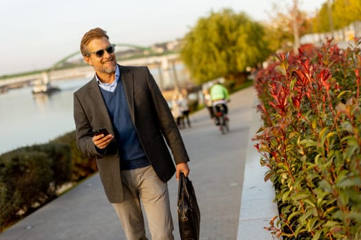 Dressed in a smart casual outfit, a man walks confidently by the water, engaged in a phone conversation while admiring the vibrant autumn colors around him