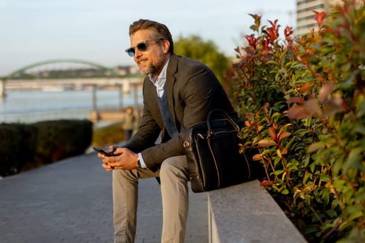 A relaxed man dressed in a stylish jacket and sunglasses sits on a stone bench by the river, engaging with his smartphone as the sun sets behind him, casting warm colors over the scene.