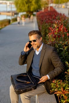 A professional man dressed in a blazer enjoys a sunny afternoon, chatting on his phone beside lush plants while carrying a leather bag. The scene reflects a blend of work and relaxation.