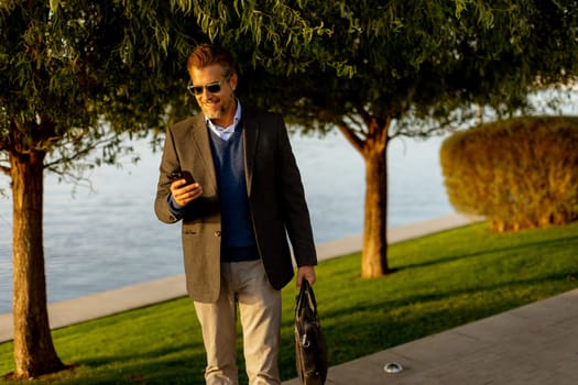 Dressed in a smart casual outfit, a man walks confidently by the water, engaged in a phone conversation while admiring the vibrant autumn colors around him