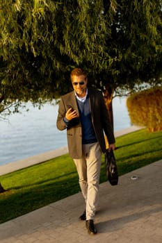 Dressed in a smart casual outfit, a man walks confidently by the water, engaged in a phone conversation while admiring the vibrant autumn colors around him
