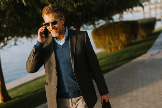 A well-dressed man strolls by the water, absorbed in a phone call. Surrounded by lush greenery, he pairs sophistication with casual ease on a sunny day.