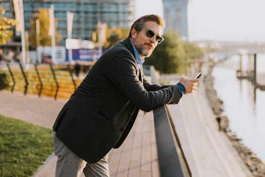 A relaxed man leans on a railing, enjoying a phone conversation as the sun sets over the water, casting a warm glow over the serene riverside surroundings.