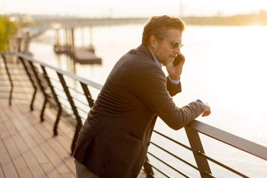 A relaxed man leans on a railing, enjoying a phone conversation as the sun sets over the water, casting a warm glow over the serene riverside surroundings.