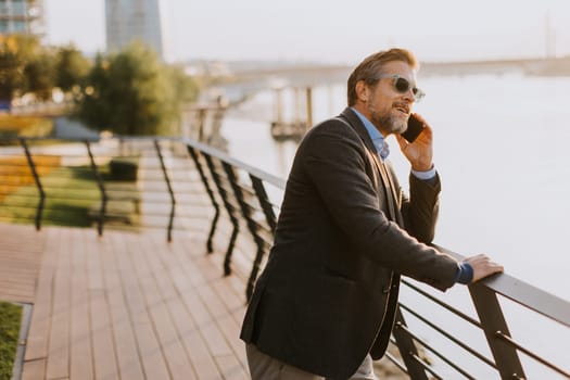 A relaxed man leans on a railing, enjoying a phone conversation as the sun sets over the water, casting a warm glow over the serene riverside surroundings.