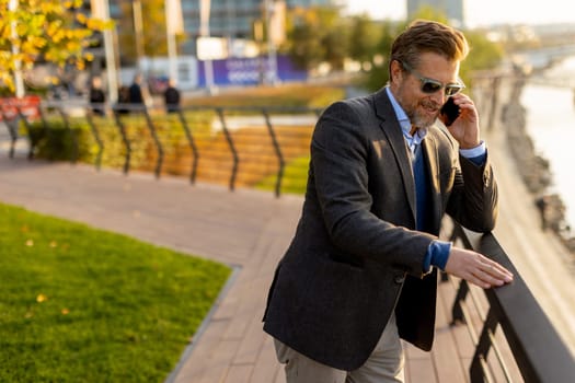 Dressed in smart casual attire, a man stands by an urban landscape, engaging in a phone conversation as autumn leaves swirl around him in the warm sunlight.