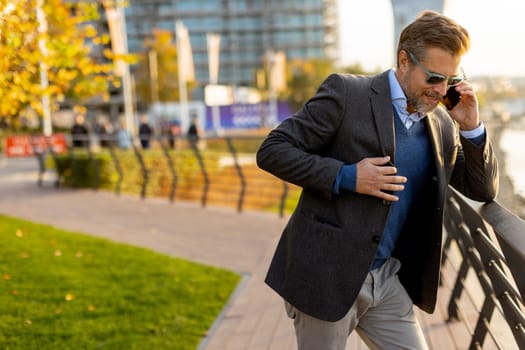 Dressed in smart casual attire, a man stands by an urban landscape, engaging in a phone conversation as autumn leaves swirl around him in the warm sunlight.