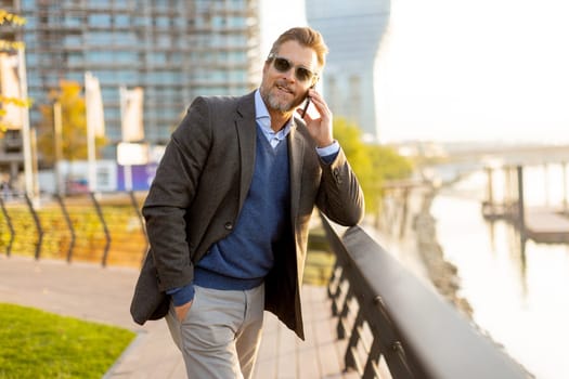Dressed in smart casual attire, a man stands by an urban landscape, engaging in a phone conversation as autumn leaves swirl around him in the warm sunlight.