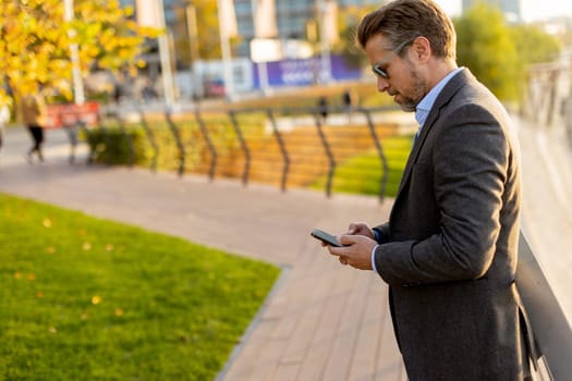 Dressed in smart casual attire, a man stands by an urban landscape, engaging in a phone conversation as autumn leaves swirl around him in the warm sunlight.