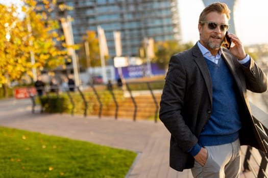 Dressed in smart casual attire, a man stands by an urban landscape, engaging in a phone conversation as autumn leaves swirl around him in the warm sunlight.