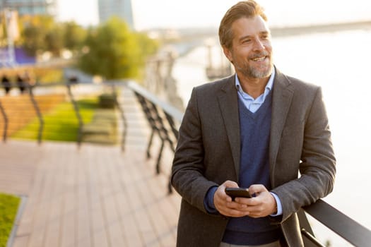 A well-dressed man takes a leisurely walk along the riverside park, engaging with his smartphone as golden hour light casts a warm glow over the landscape.