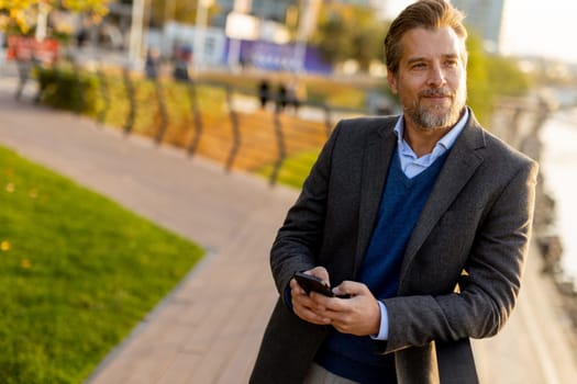 A well-dressed man takes a leisurely walk along the riverside park, engaging with his smartphone as golden hour light casts a warm glow over the landscape.