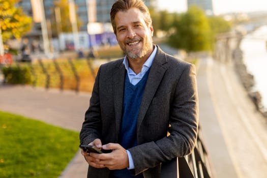 A well-dressed man takes a leisurely walk along the riverside park, engaging with his smartphone as golden hour light casts a warm glow over the landscape.