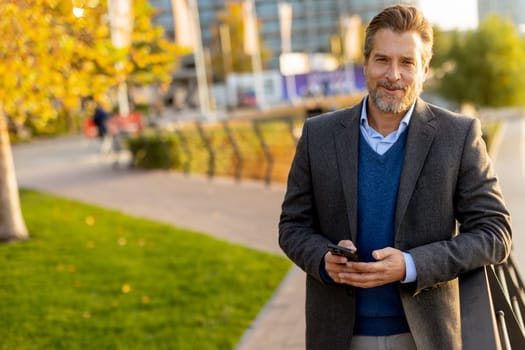 Under the warm sunlight, a relaxed professional is casually dressed, engaging with his smartphone amidst a lively outdoor environment filled with greenery and city life.