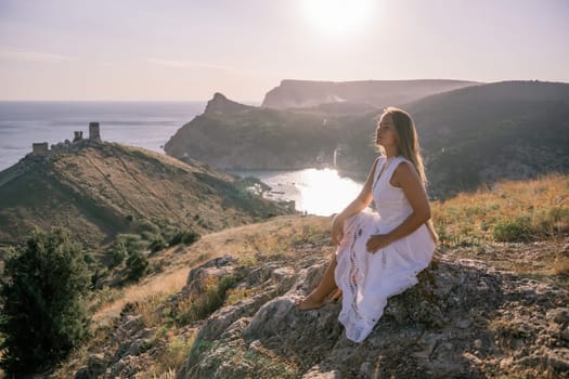 A woman in a white dress sits on a rock overlooking a body of water. The scene is serene and peaceful, with the woman enjoying the view and the calmness of the surroundings