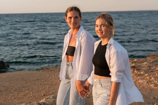 Two women, holding hands, walk along a rocky beach. They are wearing casual clothing and smiling. The ocean is visible behind them. The sky is a soft, golden color.