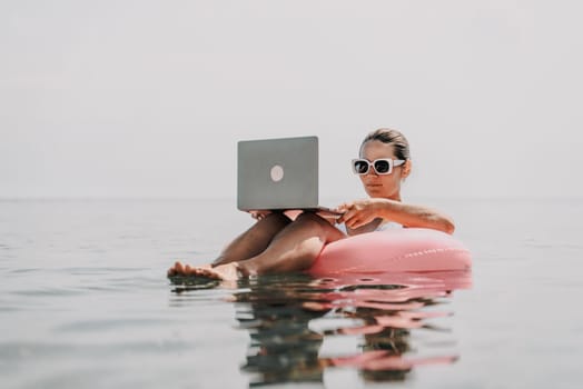 A woman is sitting on a pink inflatable raft in the ocean, using a laptop. Concept of relaxation and leisure, as the woman is enjoying her time in the water while working on her laptop
