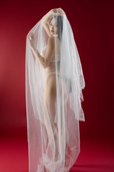 A stunning woman poses gracefully, wearing lingerie and a flowing veil in a captivating red setting.