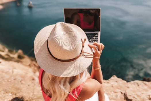 A woman is sitting on a rocky beach with a laptop in front of her. She is wearing a pink top and a straw hat. Concept of relaxation and leisure