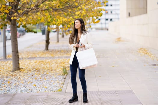 A stylish young woman walks with poise and grace down a beautiful, picturesque street adorned with colorful autumn leaves, joyfully holding a shopping bag as she enjoys her day out exploring