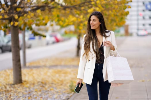 A fashionable woman enjoys a delightful autumn shopping day, confidently holding colorful bags in one hand and a smartphone in the other, savoring every joyful moment of her outing