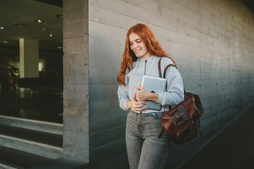 A fashionable young woman confidently holds her laptop while strolling through a bustling urban setting, showcasing the perfect blend of modern technology and stylish urban fashion in her daily life