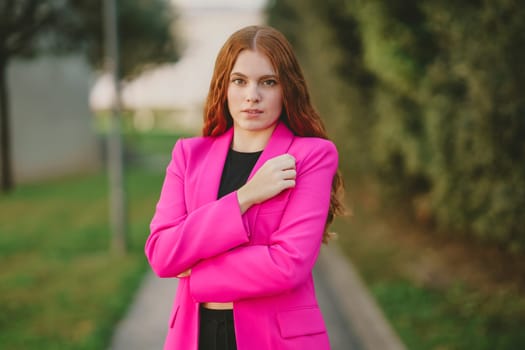In an outdoor setting that showcases lush greenery and vibrant colors, a stylish young woman, dressed in a striking pink blazer, poses confidently, exuding an air of elegance and modern femininity