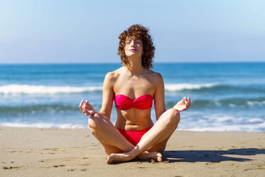 Full body of young curly ginger haired female, in pink bikini sitting in meditation with crossed legs and eyes closed while fingers in mudras in sunlight against foamy rippling seawater