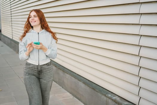 A cheerful young woman with curly red hair is outdoors, checking her smartphone in stylish casual wear. Modern architecture enhances the vibrant atmosphere of the lively urban environment around her
