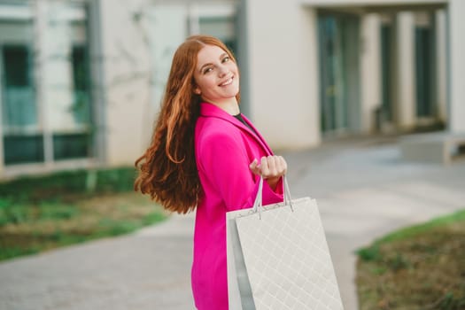 A stylish woman dressed in a vibrant bright pink blazer joyfully carries a shopping bag while outdoors, perfectly embodying the essence of fashion, sophistication, and pure happiness