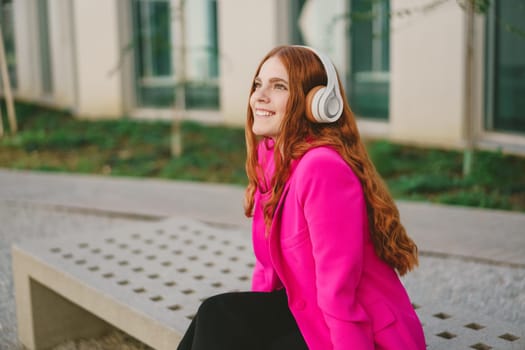 On a beautiful day, a vibrant young woman with long red hair enjoys music outdoors. Wearing stylish headphones and a bright pink jacket, she embodies carefree enjoyment and a casual lifestyle